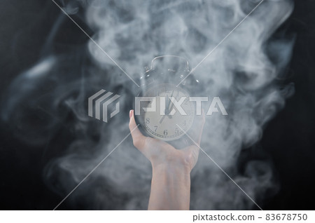 Close-up of a female hand holding a clock on a black background in smoke. Alarm clock at midnight in a mystical fog. 83678750
