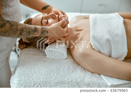 Therapist is doing face relaxing massage to a young woman in modern wellness cabinet 83679965