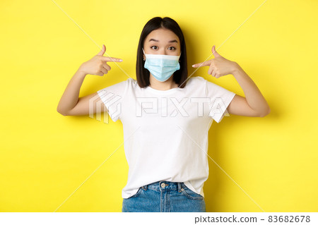 Covid-19, quarantine and social distancing concept. Young asian woman in white t-shirt, pointing at her medical mask from coronavirus, yellow background 83682678