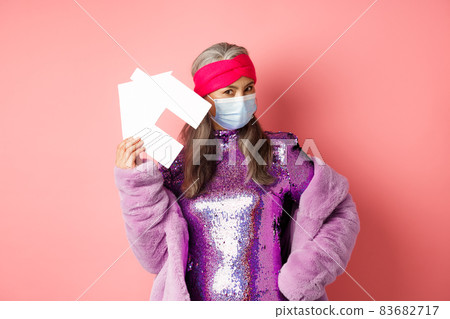 Covid-19 and real estate concept. Cool and stylish asian elderly woman in disco dress and medical mask showing paper house, standing over pink background 83682717