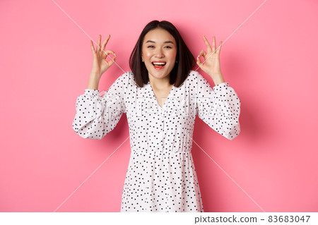 Pretty young asian woman in dress showing okay sign, praising and showing approval, looking satisfied, standing against pink background Pretty young asian woman in dress showing okay sign, praising and showing approval, looking satisfied, standing against pink background 83683047