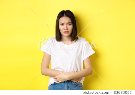 Image of asian woman feeling sick, having painful cramps, holding hands on belly and frowning from pain, discomfort during menstrual period, standing over yellow background 83683095