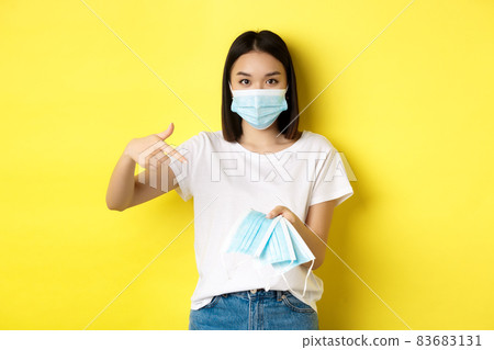 Coronavirus, quarantine and medicine concept. Young asian woman pointing finger at medical masks for going outdoors, standing over yellow background 83683131