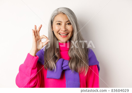 Close up of beautiful asian mature woman showing OK sign and smiling, approve and like product, standing satisfied against white background Close up of beautiful asian mature woman showing OK sign and smiling, approve and like product, standing satisfied against white background 83684029