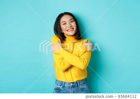 Cheerful beautiful girl embracing own body, smiling and cuddling herself, standing carefree in yellow pullover against blue background Cheerful beautiful girl embracing own body, smiling and cuddling herself, standing carefree in yellow pullover against blue background 83684112