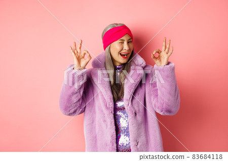 Fashion and shopping concept. Stylish asian granny saying all okay and winking. Trendy senior woman in winter fake-fur coat show OK sign, praising good product, pink background 83684118