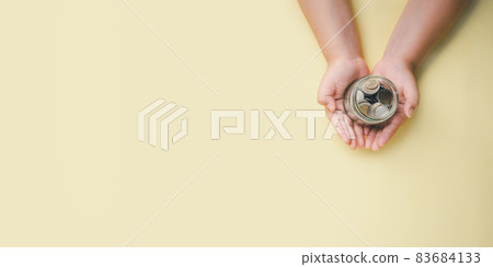 Little boy hand holding money coin in glass jar on yellow background, donation, saving, fundraising charity, family finance plan concept, superannuation, financial crisis concept, Top view. Little boy hand holding money coin in glass jar on yellow background, donation, saving, fundraising charity, family finance plan concept, superannuation, financial crisis concept, Top view. 83684133