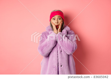 Fashion concept. Trendy old asian woman looking surprised at camera. Mature female in purple coat and headband looking amazed at camera, checking out promotion on pink background Fashion concept. Trendy old asian woman looking surprised at camera. Mature female in purple coat and headband looking amazed at camera, checking out promotion on pink background 83685042