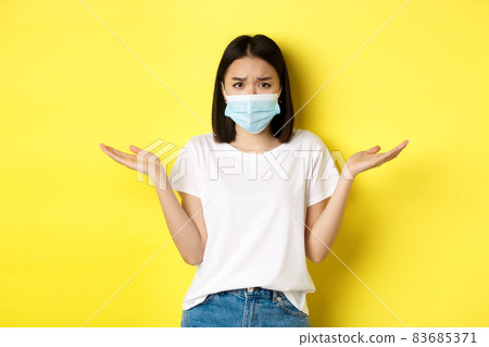 Covid-19, quarantine and social distancing concept. Confused asian woman in medical mask shrugging shoulders, spread hands sideways clueless, know nothing, yellow background 83685371