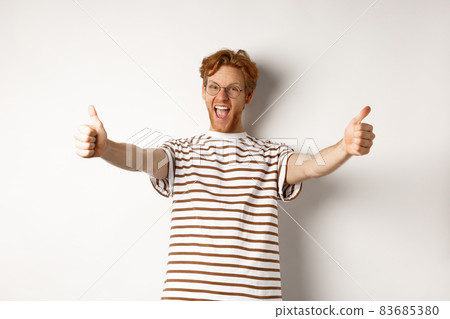 Happy redhead man scream yes and showing thumbs-up, approve and praise excellent company, standing over white background 83685380