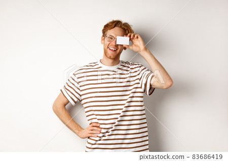 Shopping and finance concept. Happy young man with red hair and glasses showing plastic credit card near face, smiling joyfully at camera, standing over white background Shopping and finance concept. Happy young man with red hair and glasses showing plastic credit card near face, smiling joyfully at camera, standing over white background 83686419