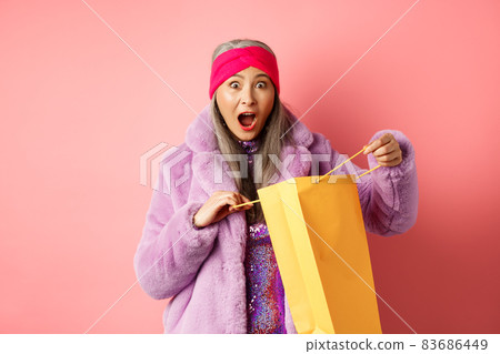 Shopping and fashion concept. Surprised asian grandmother open shop bag and looking amazed at camera, receive mother day gift, pink background 83686449