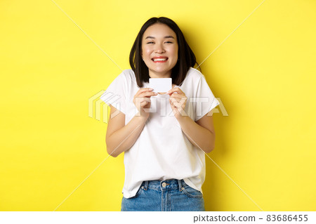 Young asian woman in casual white t-shirt going on shopping, showing plastic credit card and smiling excited, standing over yellow background Young asian woman in casual white t-shirt going on shopping, showing plastic credit card and smiling excited, standing over yellow background 83686455