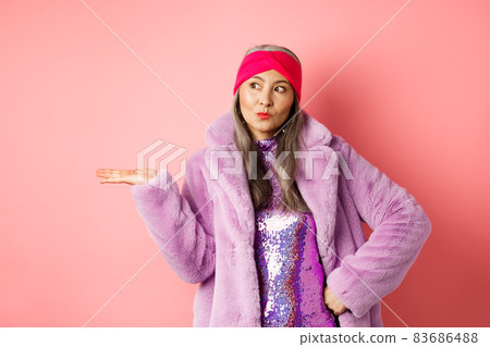 Fashion and shopping concept. Stylish asian senior woman in purple winter outfit demonstrating product on hand. Cool old lady in trendy clothes holding something on palm, pink background Fashion and shopping concept. Stylish asian senior woman in purple winter outfit demonstrating product on hand. Cool old lady in trendy clothes holding something on palm, pink background 83686488