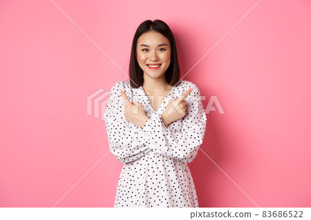 Beautiful asian girl in romantic dress pointing fingers sideways, showing two variants on shopping, smiling at camera, standing against pink background 83686522
