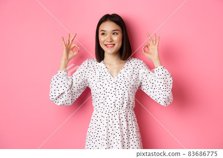 Pretty young asian woman in dress showing okay sign, praising and showing approval, looking satisfied, standing against pink background 83686775
