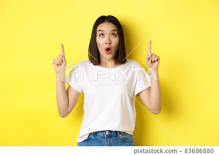 Beauty and fashion concept. Beautiful asian woman in white t-shirt pointing fingers up, standing over yellow background 83686880