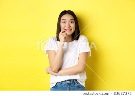 Portrait of cheerful asian girl wants something, looking with desire and temptation at camera, smiling at camera, standing ovr yellow background Portrait of cheerful asian girl wants something, looking with desire and temptation at camera, smiling at camera, standing ovr yellow background 83687375