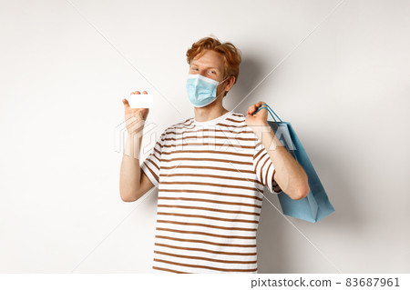Concept of covid-19 and lifestyle. Happy young shopper in face mask holding shopping bag and showing plastic credit card, buying with discounts, white background 83687961