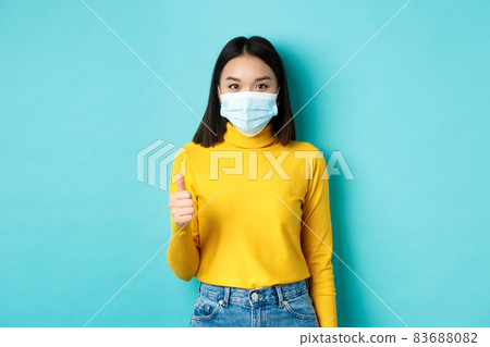 Covid-19, social distancing and pandemic concept. Young woman in medical mask showing thumbs up in approval, saying yes, standing over blue background 83688082