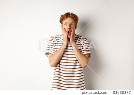 Impressed young man with ginger hair, gasping and staring shocked at camera, express complete disbelief and amazement, white background Impressed young man with ginger hair, gasping and staring shocked at camera, express complete disbelief and amazement, white background 83688377