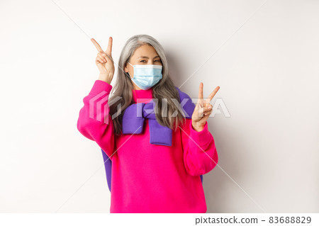 Covid, pandemic and social distancing concept. Cheerful and stylish asian senior woman wearing medical mask and showing peace signs, standing over white background 83688829
