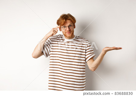 Are you stupid. Confused redhead guy in glasses shrugging, pointing at head and staring skeptical at camera, white background Are you stupid. Confused redhead guy in glasses shrugging, pointing at head and staring skeptical at camera, white background 83688830