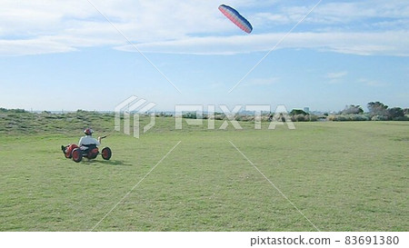 From a seaside park where a small cart on the ground advances, towed by a kite flying high in the blue sky From a seaside park where a small cart on the ground advances, towed by a kite flying high in the blue sky 83691380