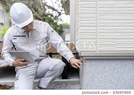A man in work clothes inspecting the foundation of a house 83695908