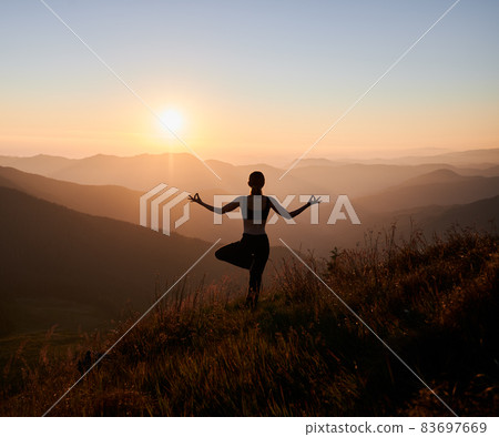Back view of slim woman performing yoga pose on grassy hill with orange sky on background. Fit woman standing on one leg and doing yoga exercise outdoors at sunset. 83697669