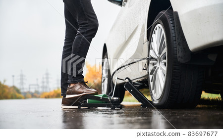 Close up of male legs in gray jeans inflating flat tire with air compressor. Young man using air compressor to inflate tire while standing by the car on rainy road. Close up of male legs in gray jeans inflating flat tire with air compressor. Young man using air compressor to inflate tire while standing by the car on rainy road. 83697677