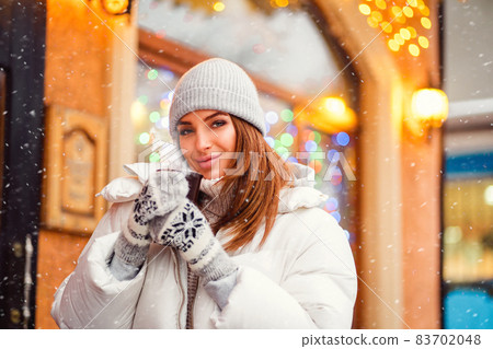 Smiling beautiful girl walks on Christmas city while holds coffe to go in her hands. Festive mood Smiling beautiful girl walks on Christmas city while holds coffe to go in her hands. Festive mood 83702048
