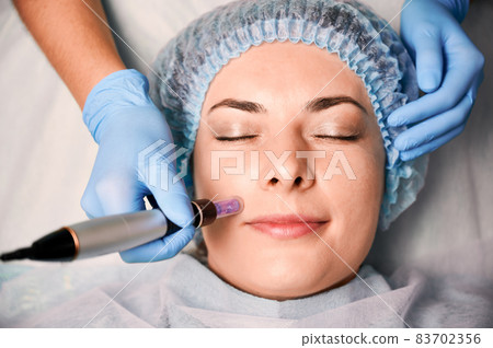 Close up of beautician hands in sterile gloves using dermapen during skincare procedure. Young woman receiving facial treatment in beauty salon. Concept of skincare and collagen induction therapy. 83702356