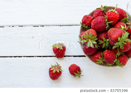 Ripe red strawberries lie in a white cup on a wooden white table. Copy space. View from above. Ripe red strawberries lie in a white cup on a wooden white table. Copy space. View from above. 83705396