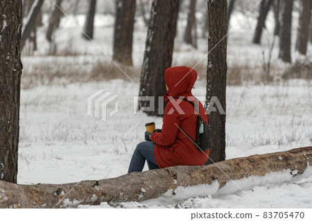 Woman in red warm clothes sits on log in winter park with cup of coffee in her hands. Walk through the winter forest. 83705470