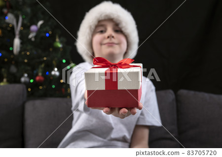 Boy in Santa hat is holding out present to you sitting on the sofa near the Christmas tree. New Year. Christmas. Boy in Santa hat is holding out present to you sitting on the sofa near the Christmas tree. New Year. Christmas. 83705720