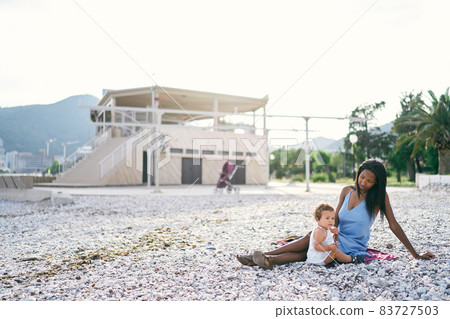 Little girl sitting next to her mother on a pebble beach against the backdrop of a building Little girl sitting next to her mother on a pebble beach against the backdrop of a building 83727503