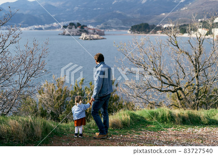 Dad holds the hand of a little girl, standing on the coast against the background of the sea, mountains and Sveti Stefan island. Back view Dad holds the hand of a little girl, standing on the coast against the background of the sea, mountains and Sveti Stefan island. Back view 83727540