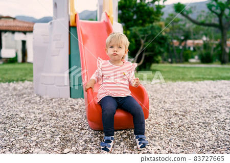 Little girl sitting on a slide in the playground Little girl sitting on a slide in the playground 83727665