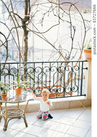 Small child in a white blouse and colorful pants sits on his knees on the balcony near a table with green flowerpots against the background of trees 83727666
