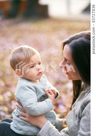 Smiling mom holding baby on her lap, face to face, sitting on foliage in the park. Close-up 83727836