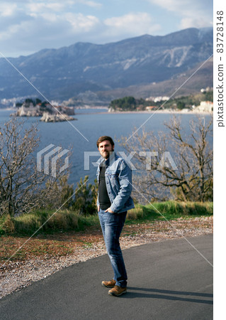 Man in a blue denim jacket and jeans stands holding his hands in his pockets on an asphalt road against the backdrop of the sea, mountains and the island of Sveti Stefan Man in a blue denim jacket and jeans stands holding his hands in his pockets on an asphalt road against the backdrop of the sea, mountains and the island of Sveti Stefan 83728148