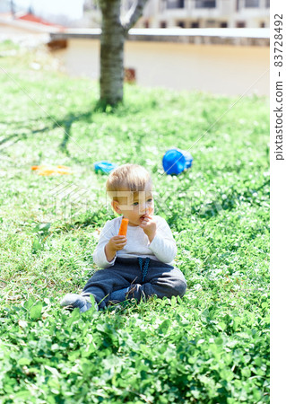 Kid sits on the green grass and gnaws a carrot on a wood background Kid sits on the green grass and gnaws a carrot on a wood background 83728492