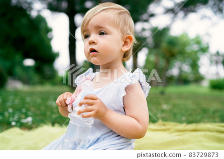 Little girl sits on a bedspread and holds a bottle of water in her hand 83729873