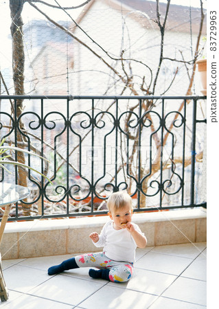Small child in a white blouse and bright pants sits on the balcony near the table and gnaws cookies on the background of trees 83729963