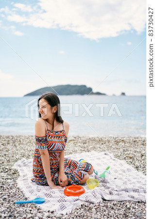 Smiling girl sitting on a blanket on a pebble beach, turning her head to the side 83730427