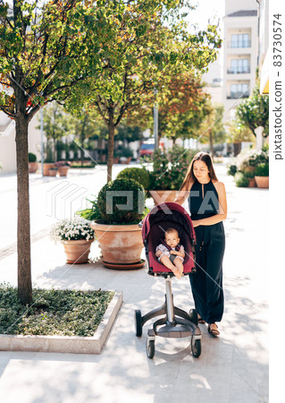Mom rolls the stroller with the baby past the tubs with green bushes Mom rolls the stroller with the baby past the tubs with green bushes 83730574