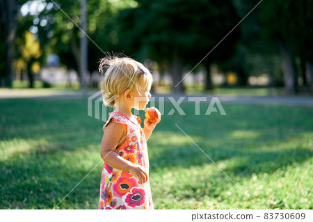 Little girl stands with a peach on a green lawn. Side view 83730609