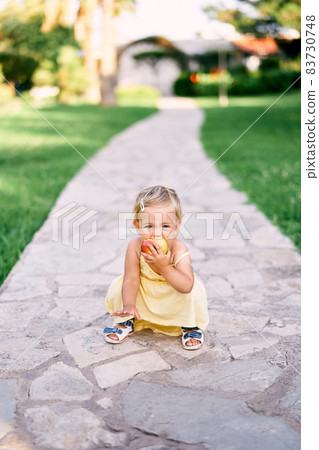 Little girl squatting on a paved path in the park and nibbling an apple Little girl squatting on a paved path in the park and nibbling an apple 83730748