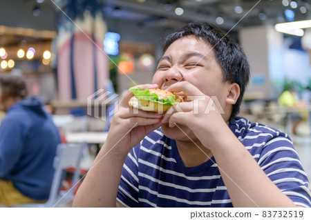 Obese boy eats a hamburger in a food court in a shopping center. junk food 83732519